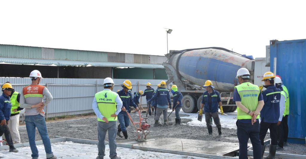 Nhóm công nhân đội mũ bảo hộ và mặc áo phản quang trao đổi tại công trường có xe trộn bê tông.
