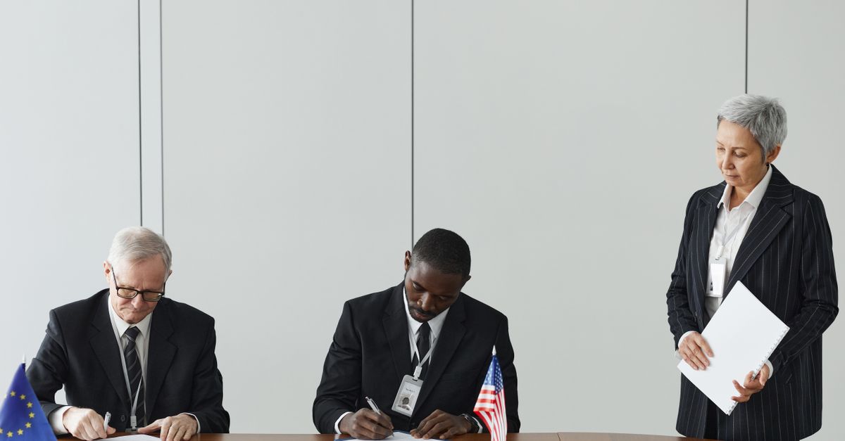 Executives signing international agreement with EU and US flags displayed on a wooden table.
