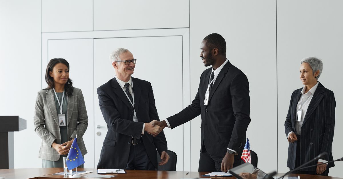 Business professionals shaking hands in conference room with flags signifying international agreement.