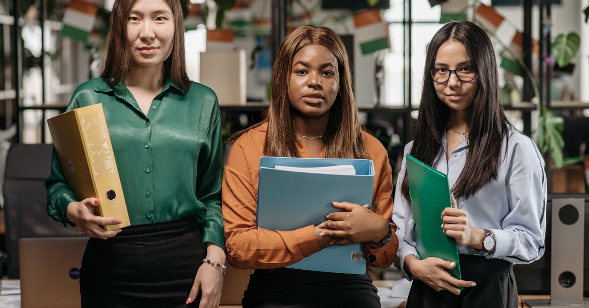 Three diverse women holding binders in an office, representing teamwork and diversity.