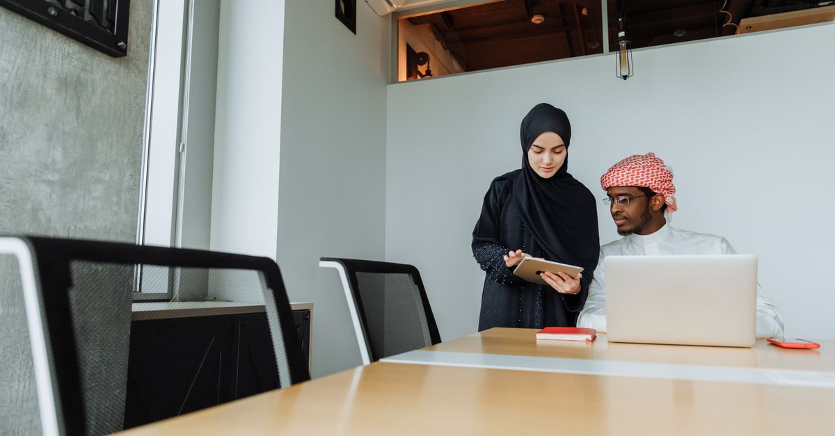 A man and woman in traditional clothing collaborate in a modern office setting, using tablets and laptops.