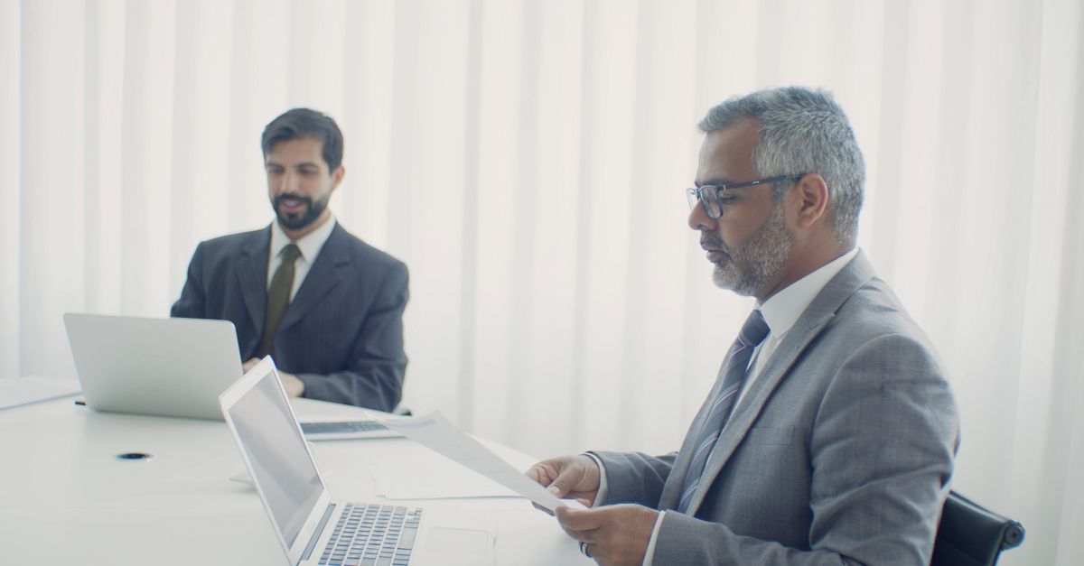 Two colleagues in a corporate setting, working on laptops and documents.